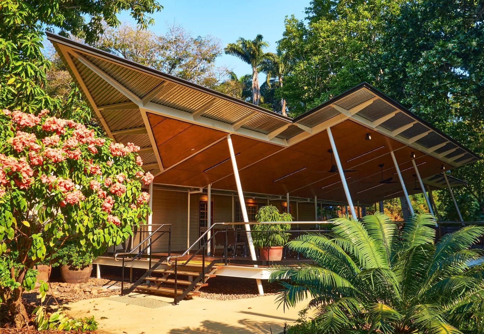 Modern house with a sharp, angular roof and wooden ceiling set in a lush, sunlit tropical garden.
