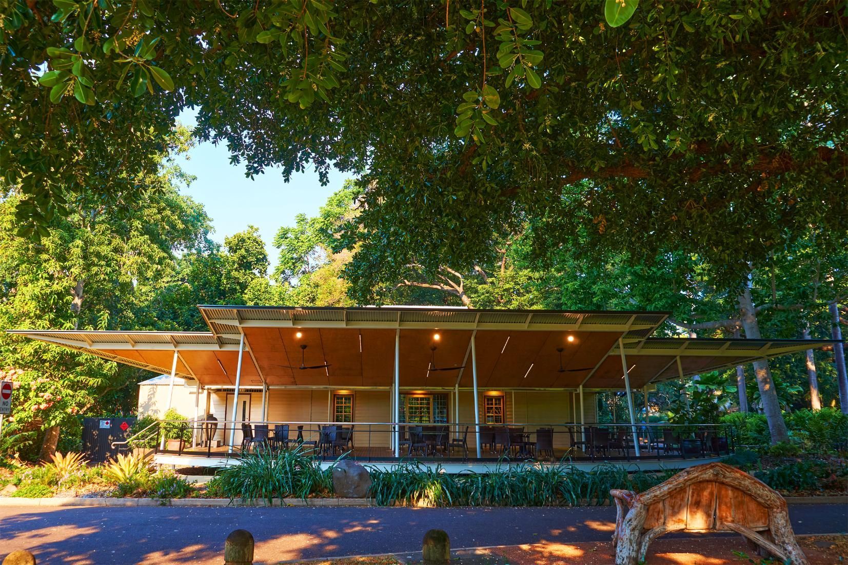 An open-air pavilion with wooden ceilings and outdoor seating, nestled under a canopy of lush green trees in a park.