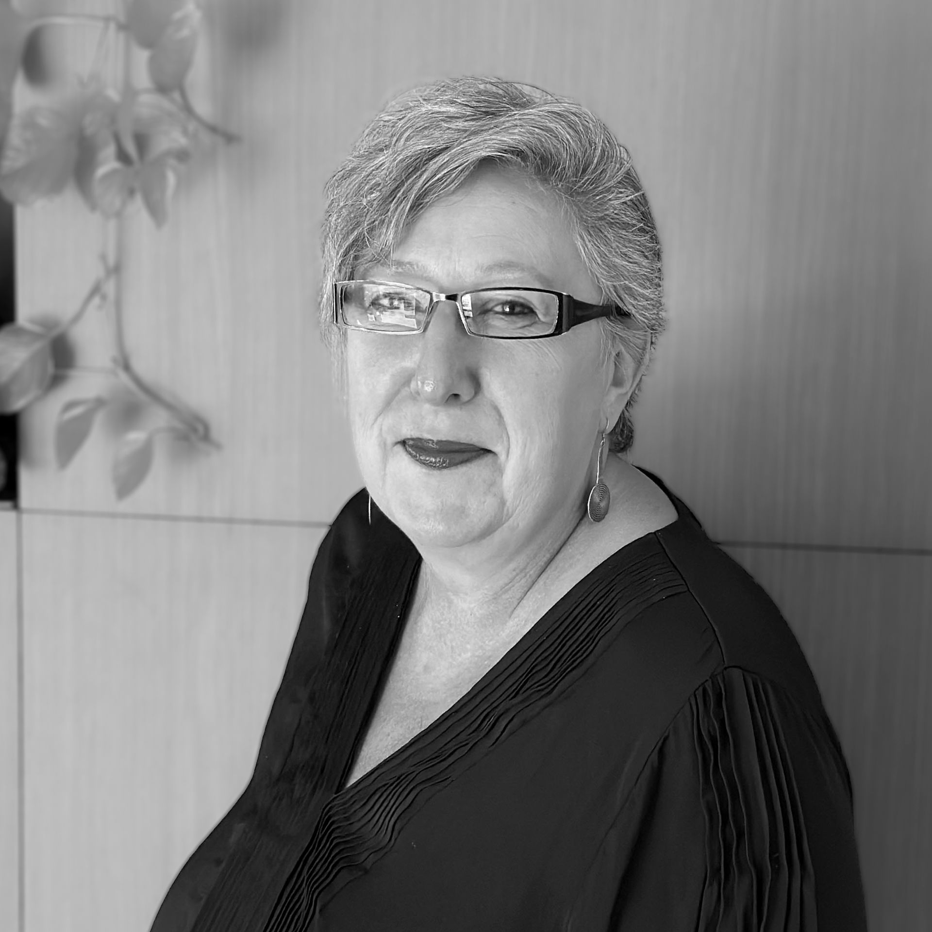 A person with short hair and glasses wearing a dark, textured blouse, captured in a portrait against a paneled wall.