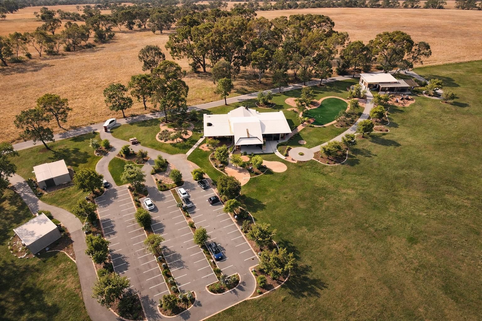 An aerial view of a rural facility with a white main building, parking lot, landscaped gardens, and surrounding fields.