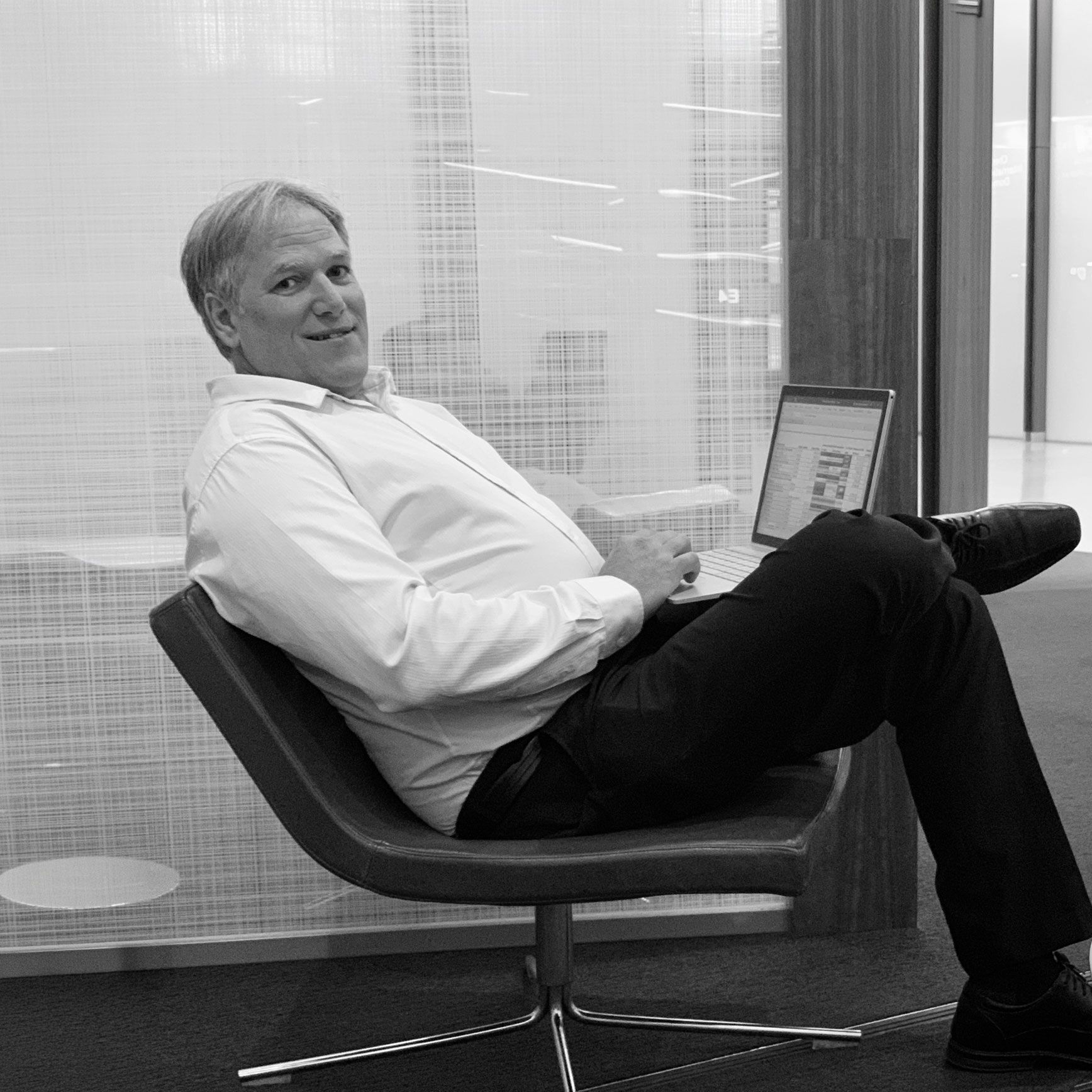 A person sitting in a lounge chair with legs crossed, typing on a laptop in a brightly lit office setting.