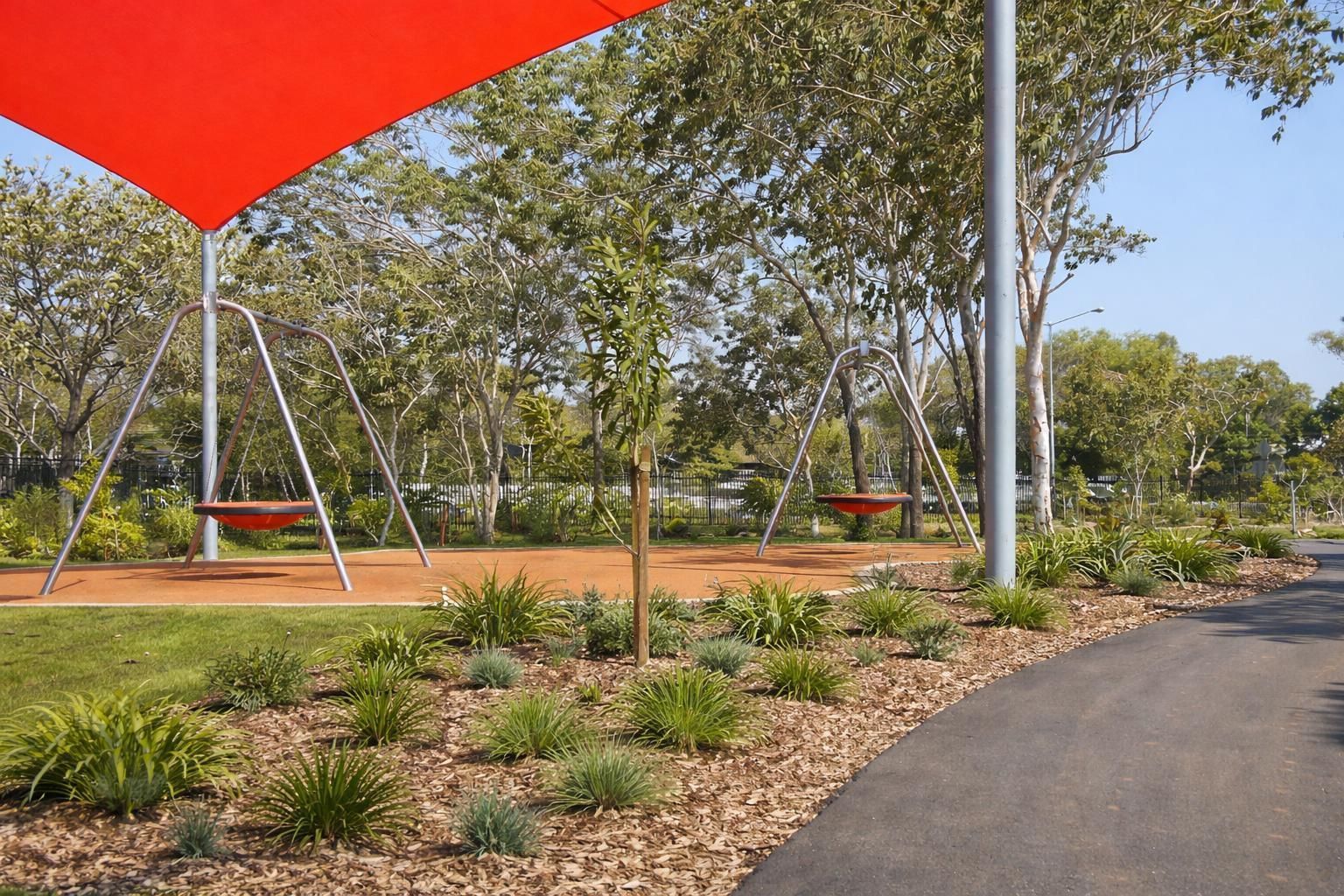 A playground with two saucer swings under a red shade sail, surrounded by garden beds, trees, and a paved walkway.