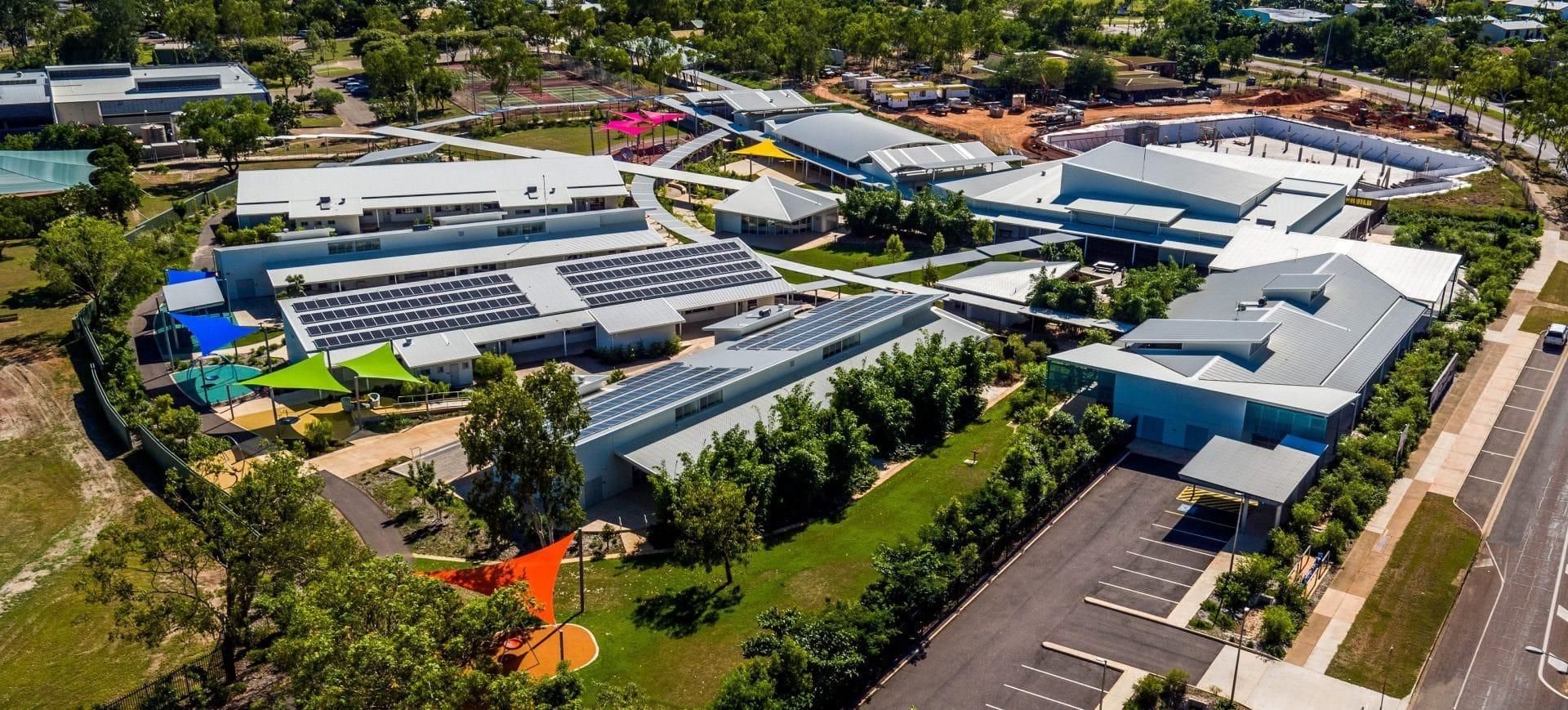Aerial view of a campus with multiple buildings, solar panels, green landscaping, parking areas, and trees.