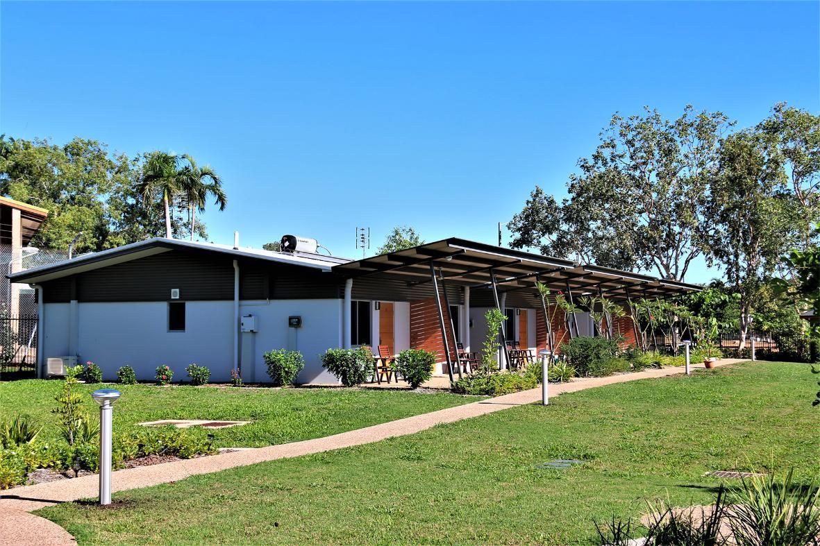 A single-story building with a dark roof and white walls sits beside a grassy lawn and a walkway under a wooden pergola.