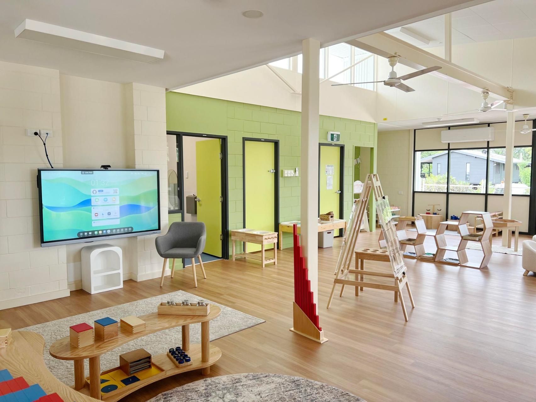 An indoor learning space with a digital whiteboard, wooden tables, chairs, and play equipment on a light wood floor.