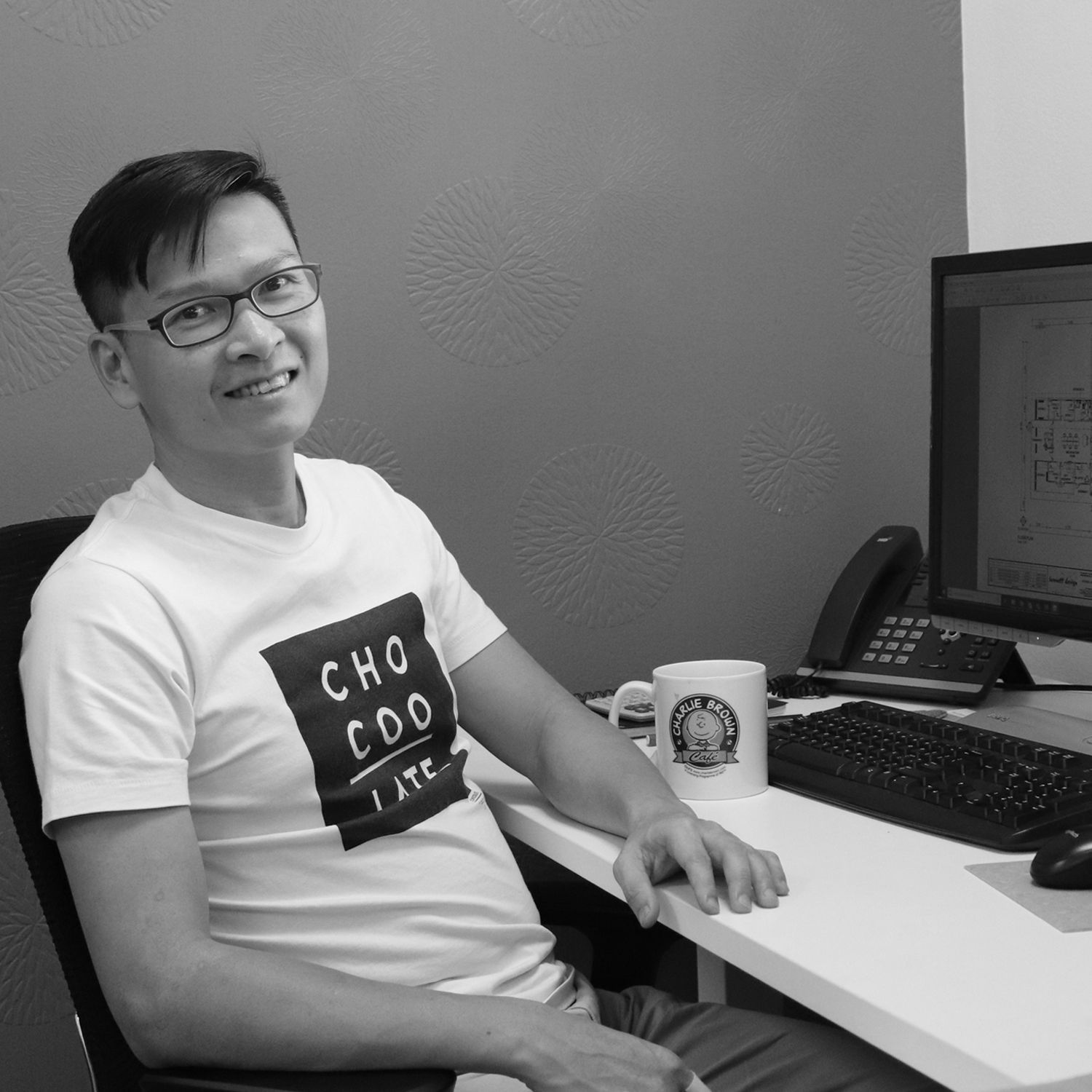 A smiling professional sits at a desk with a computer and mug, wearing a t-shirt with the word 
