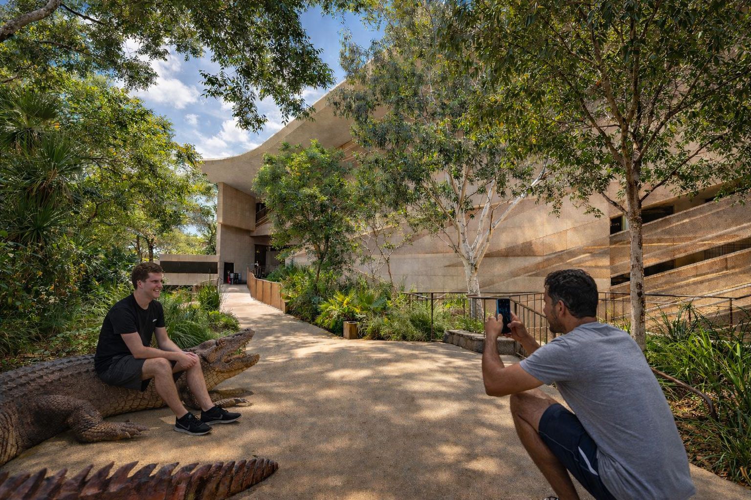 A person photographs another sitting on a large crocodile sculpture on a garden path near a modern building.