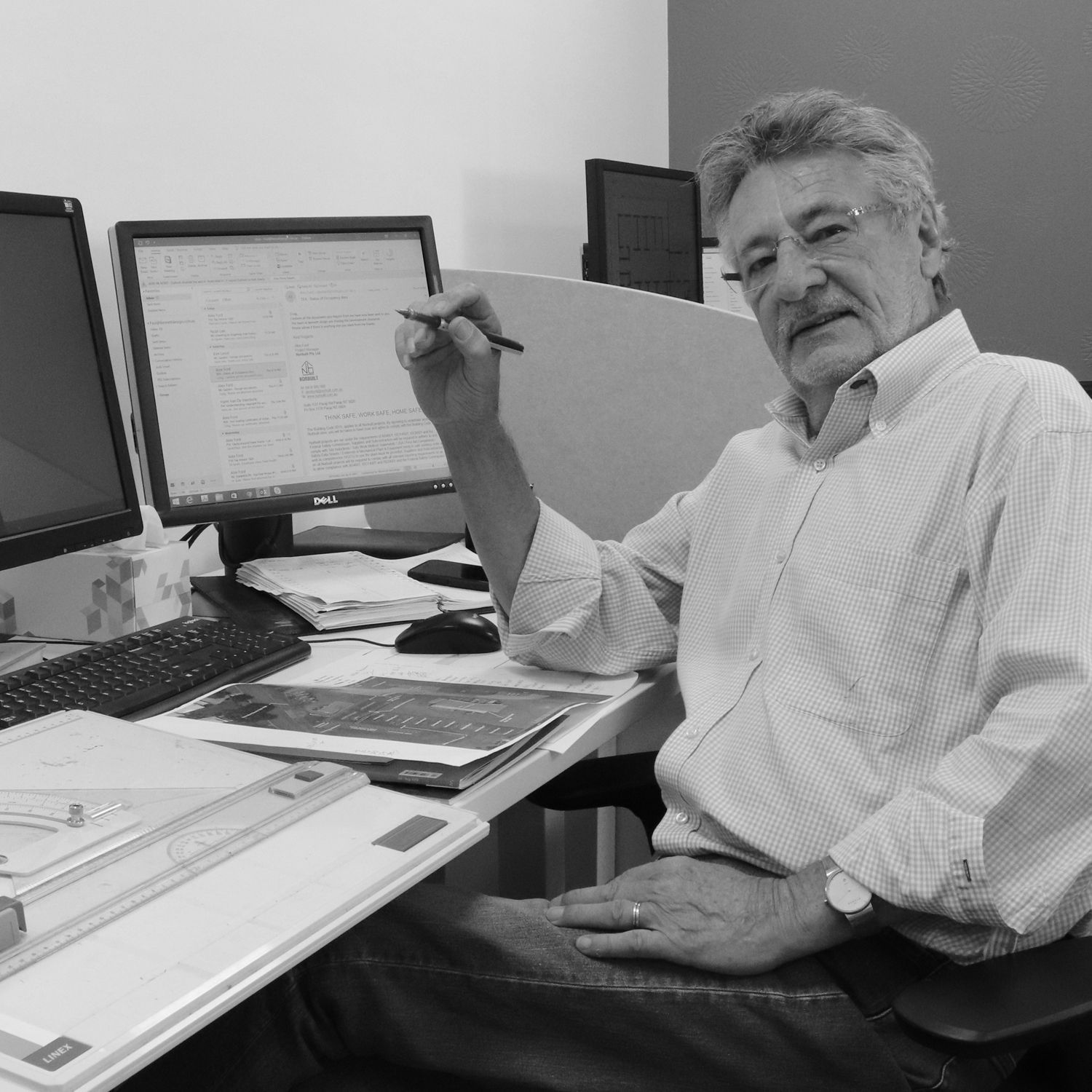 A person sits at a desk with two computer monitors, holding a pen and looking toward the camera in an office setting.