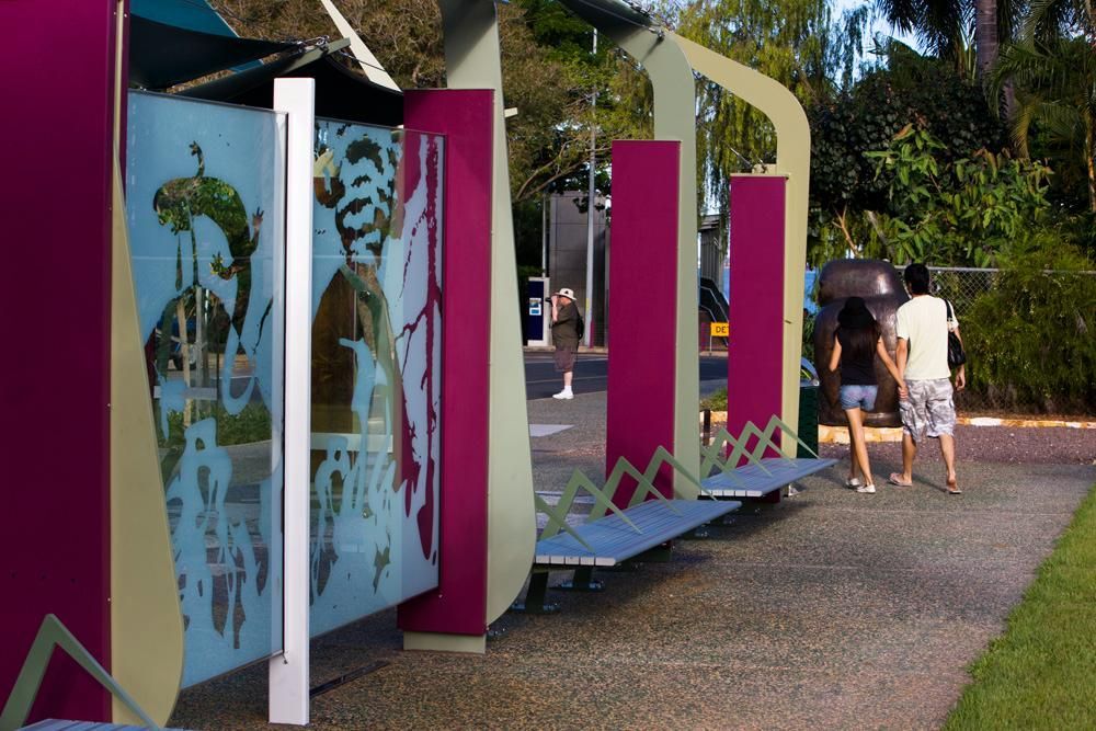A public outdoor walkway with decorative glass panels, magenta columns, metal benches, and people walking nearby.