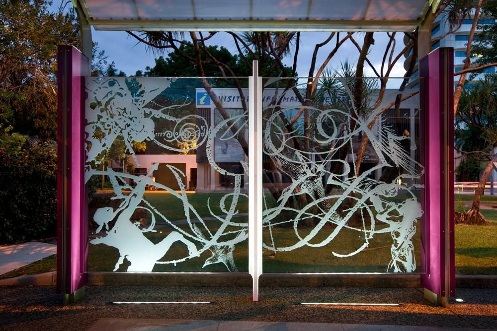 A modern bus shelter with glass panels featuring white, swirling artistic designs, illuminated by purple side lights.