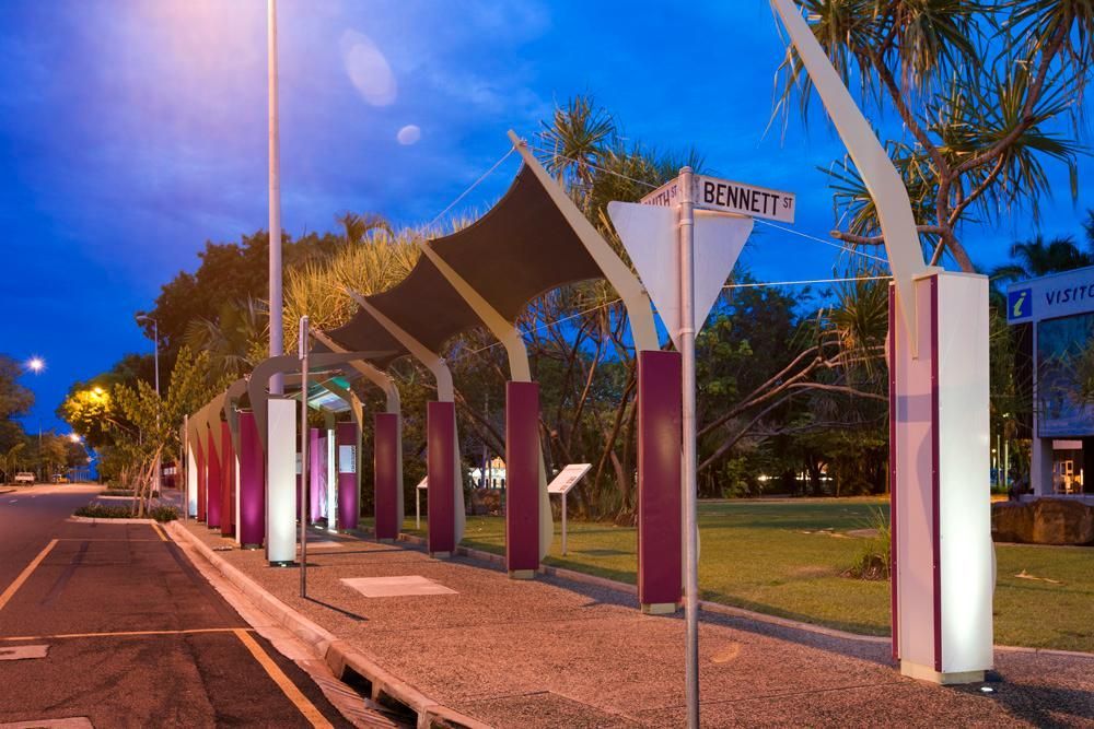 A modern bus stop with purple pillars and a dark shade canopy, illuminated at twilight beside a street and green lawn.