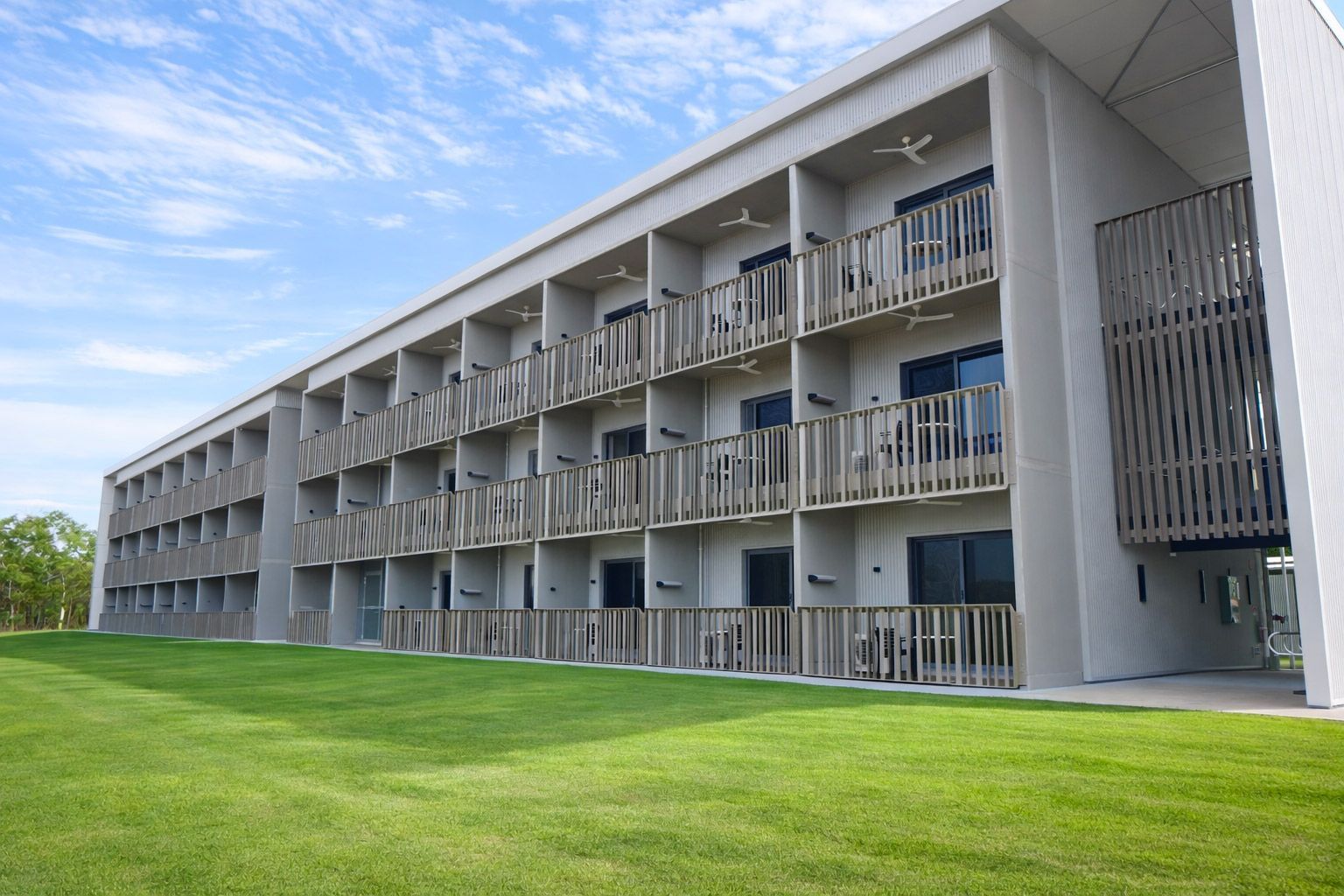 A multi-story hotel building with gray concrete walls and wooden slatted balcony railings, set against a green lawn.