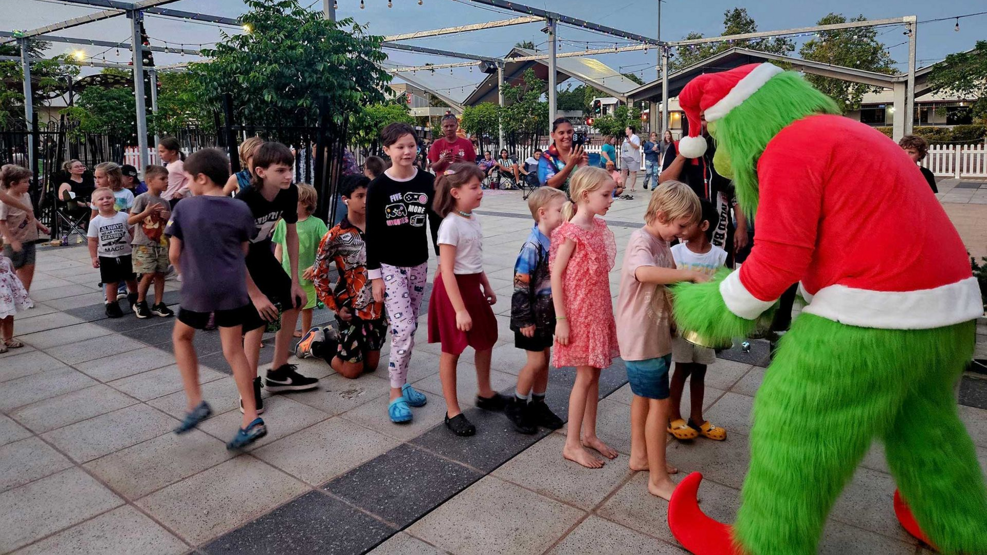 A mascot dressed as the Grinch in a Santa suit interacts with children in a sunny outdoor plaza.