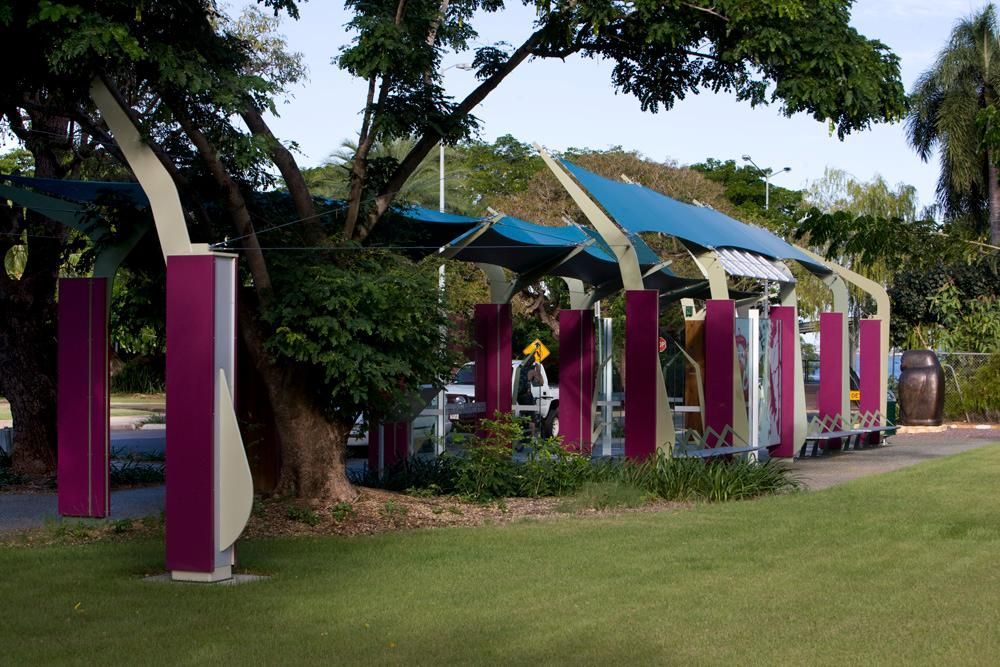 A public shelter with purple columns and blue fabric sails, set in a sunny park with trees and a green lawn.