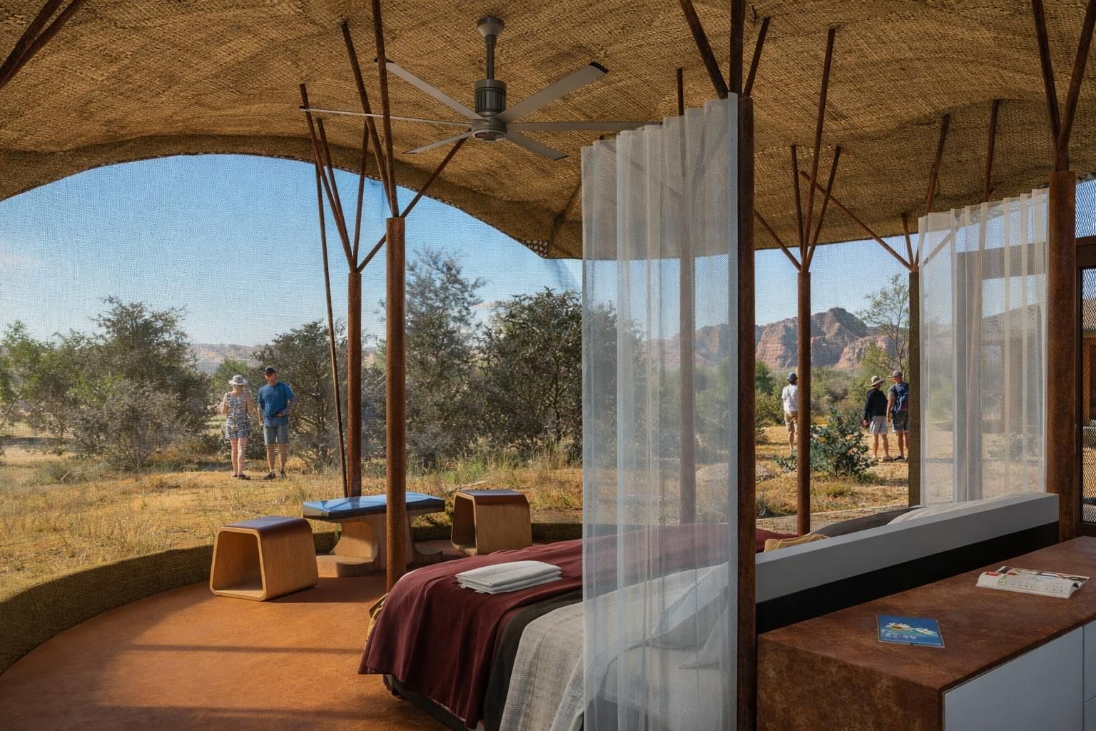 A desert safari tent with an open-air front, a bed with a white canopy, wooden stools, and views of a dry, shrubby landscape.