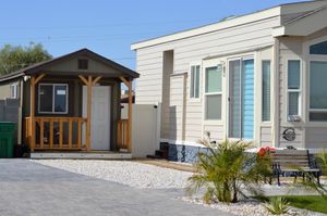 Small porch cabin beside a white mobile home on a gravel lot, with patio chairs and potted plants.