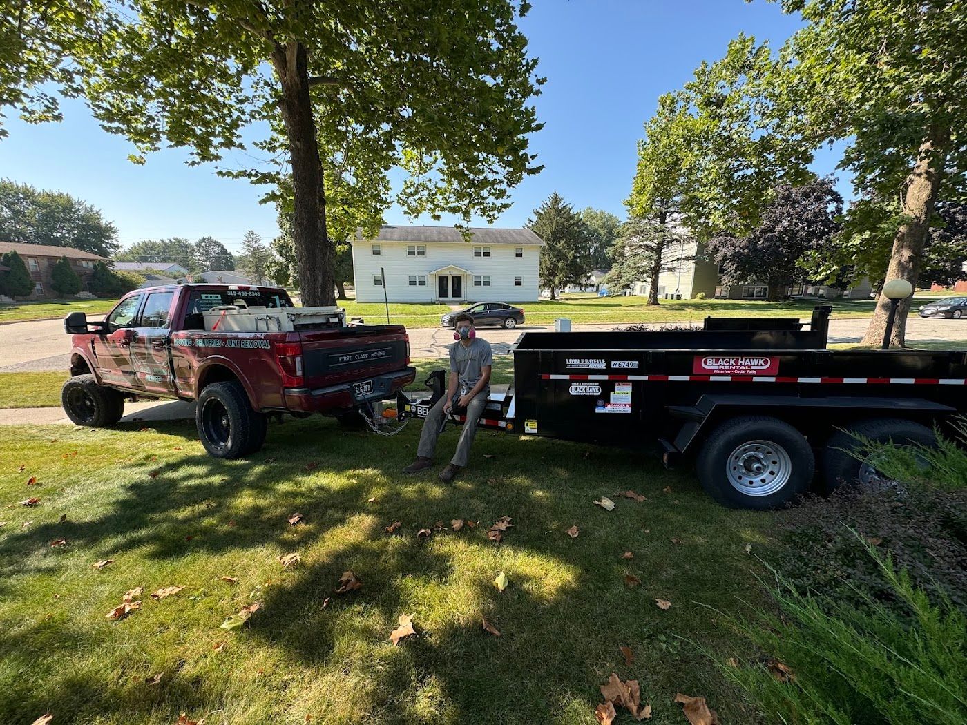 Red truck towing a black trailer with a man standing nearby on a grassy lawn under a tree.