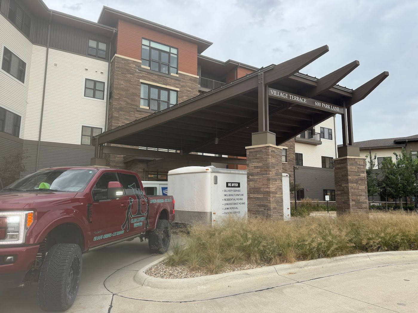 A maroon pickup truck pulling up to a building entrance with a brown pergola. A white container sits nearby.