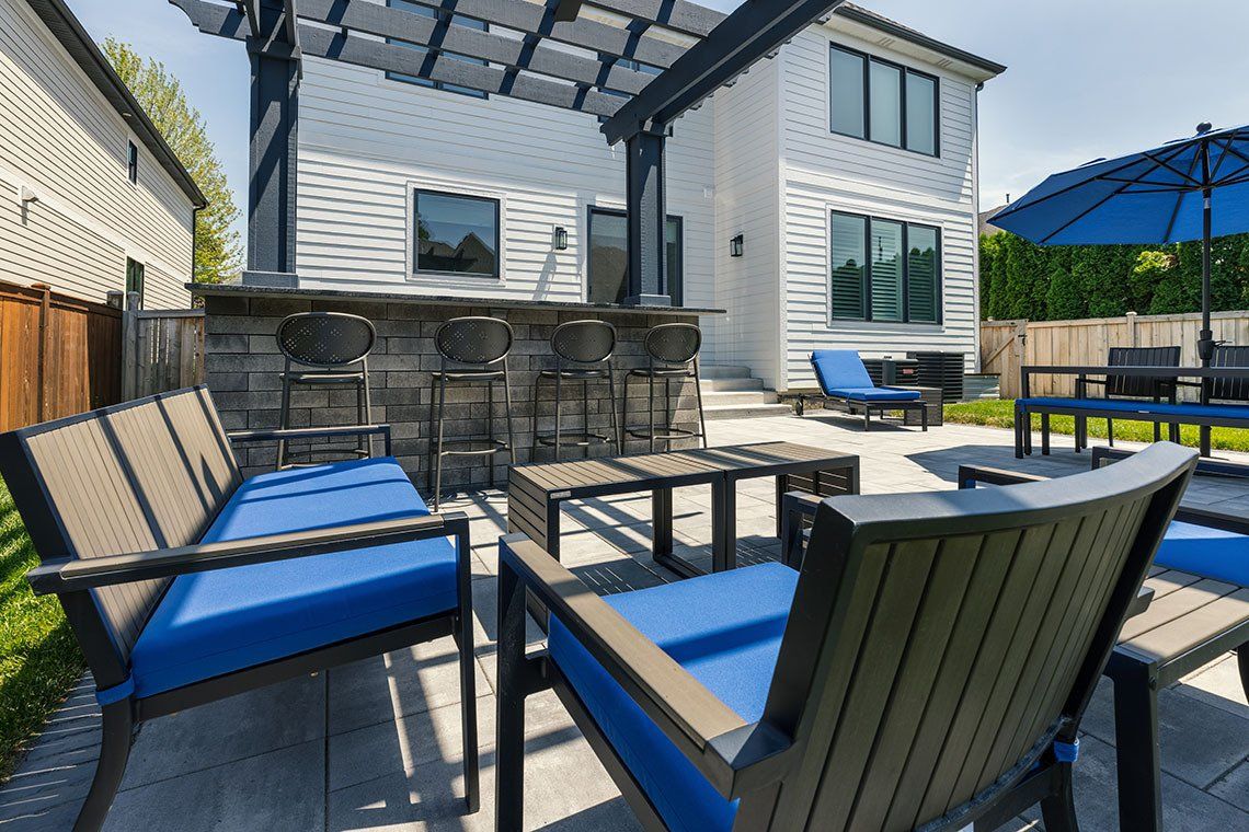 A patio with a couch , chairs , tables and umbrellas in front of a house.