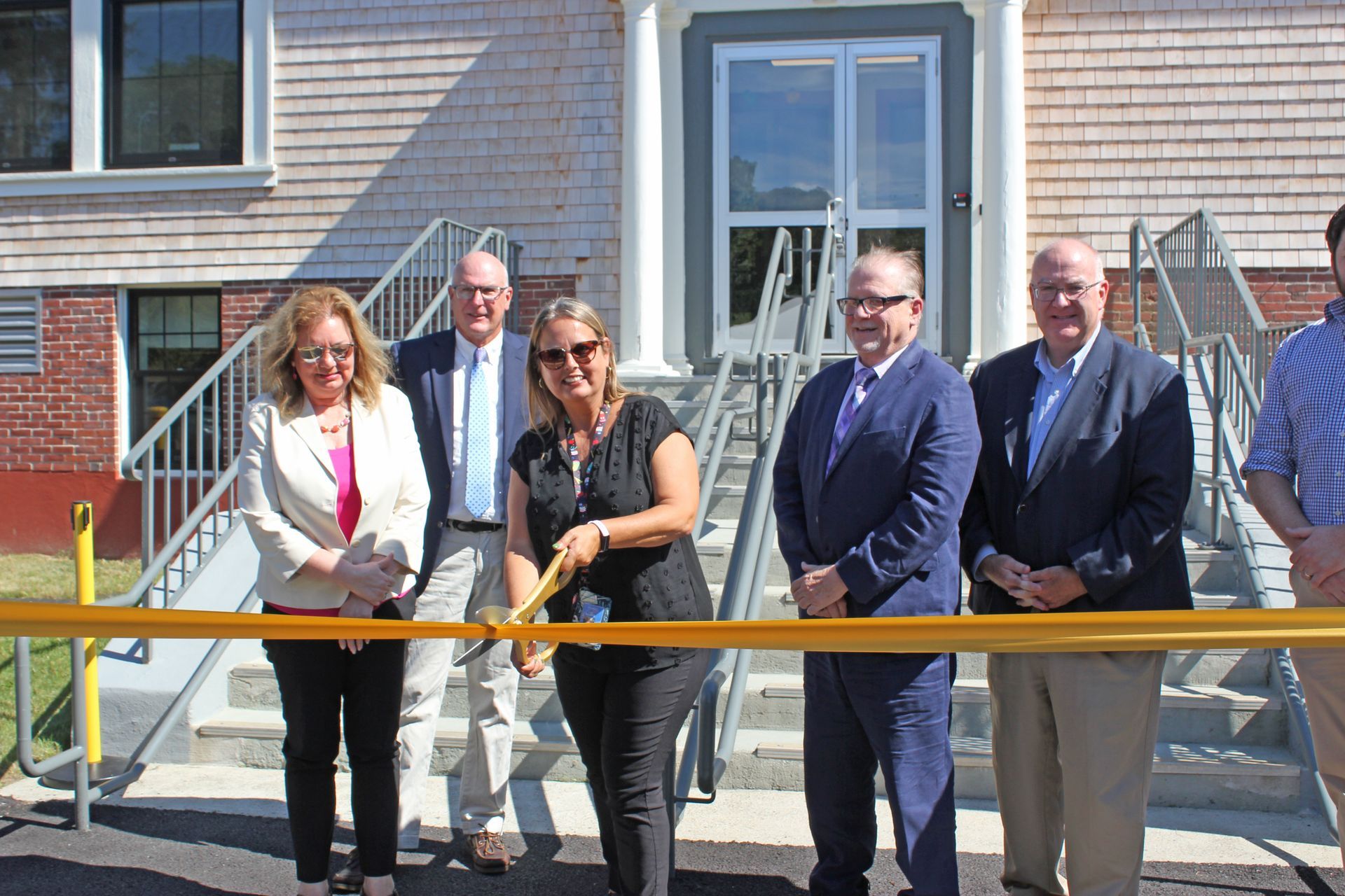 Group cuts gold ribbon in front of building; sunny day.