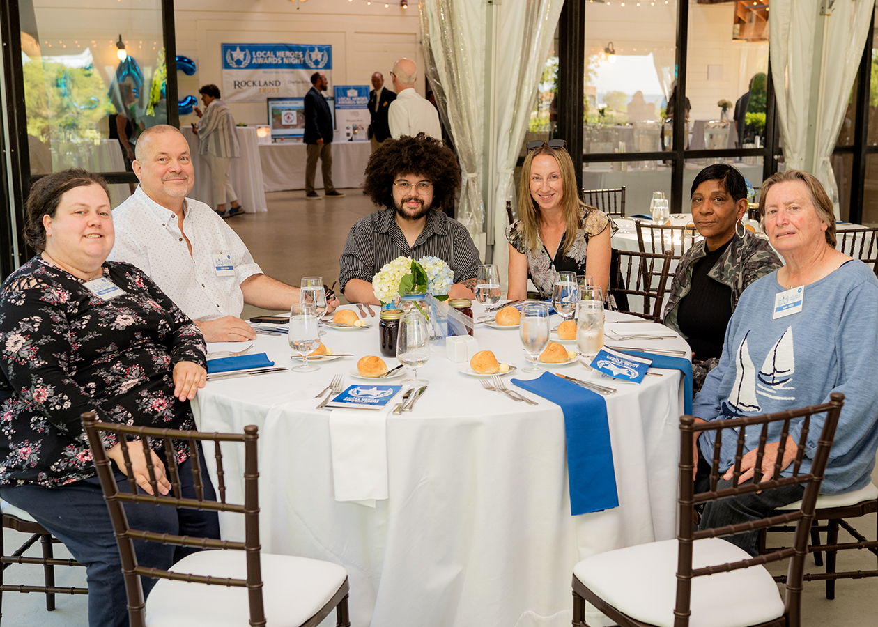 Five people seated at a round table in a decorated hall, smiling and eating at an event
