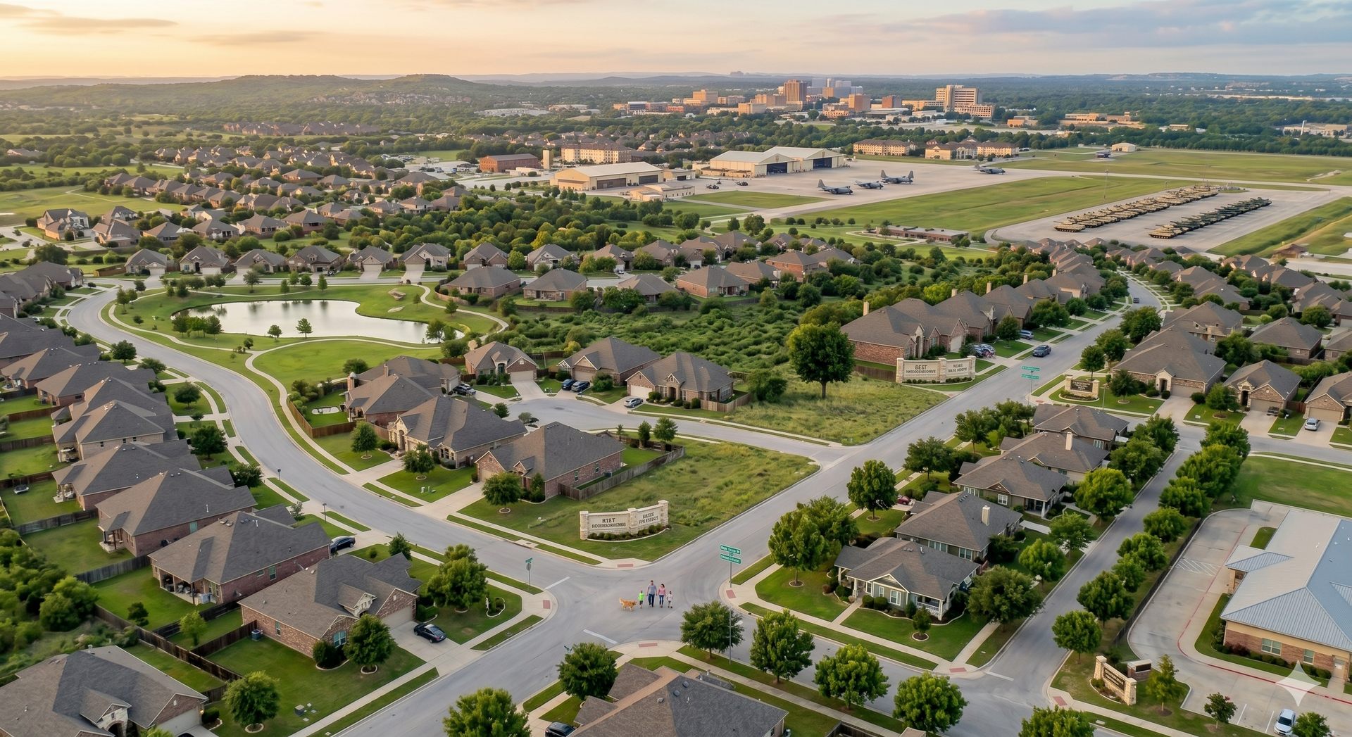 Aerial view of a suburban neighborhood with curving streets, houses, ponds, and a sunset skyline in the distance