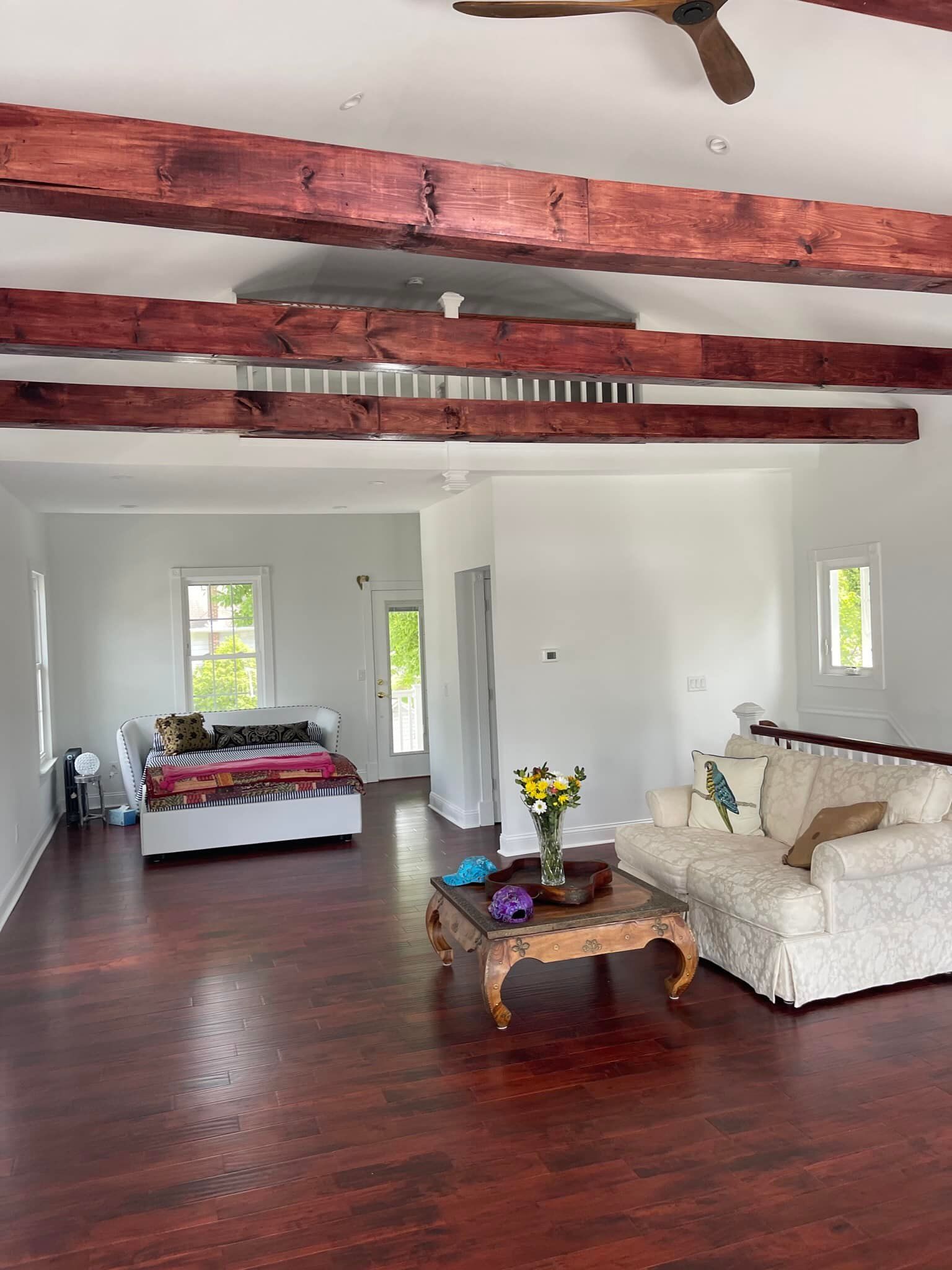 Interior with dark wood floor, white walls, and exposed dark wood beams. Sofas, table, bed, and flowers.