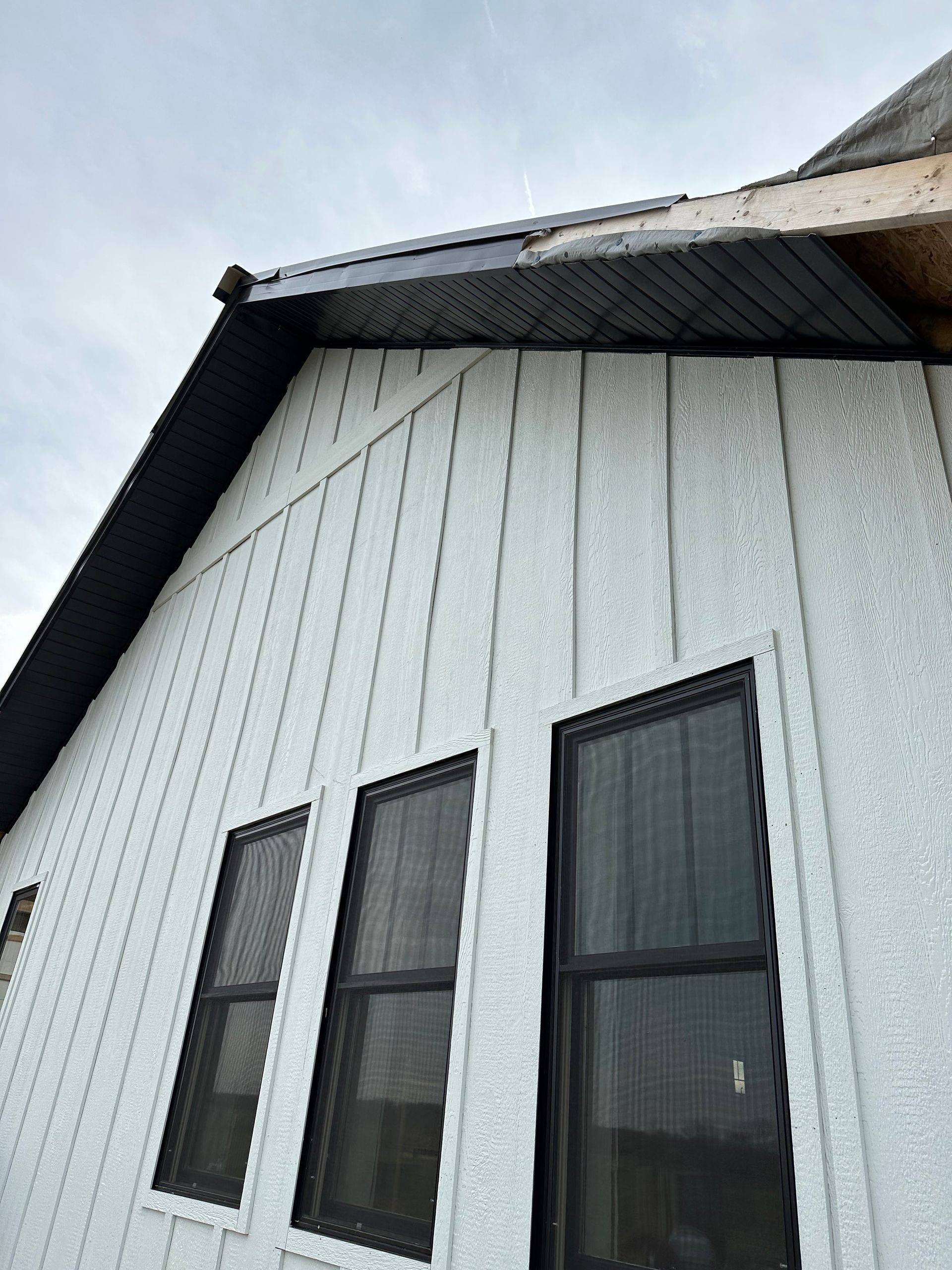 Exterior of a building with white vertical siding and three black-framed windows. The roof has black trim.