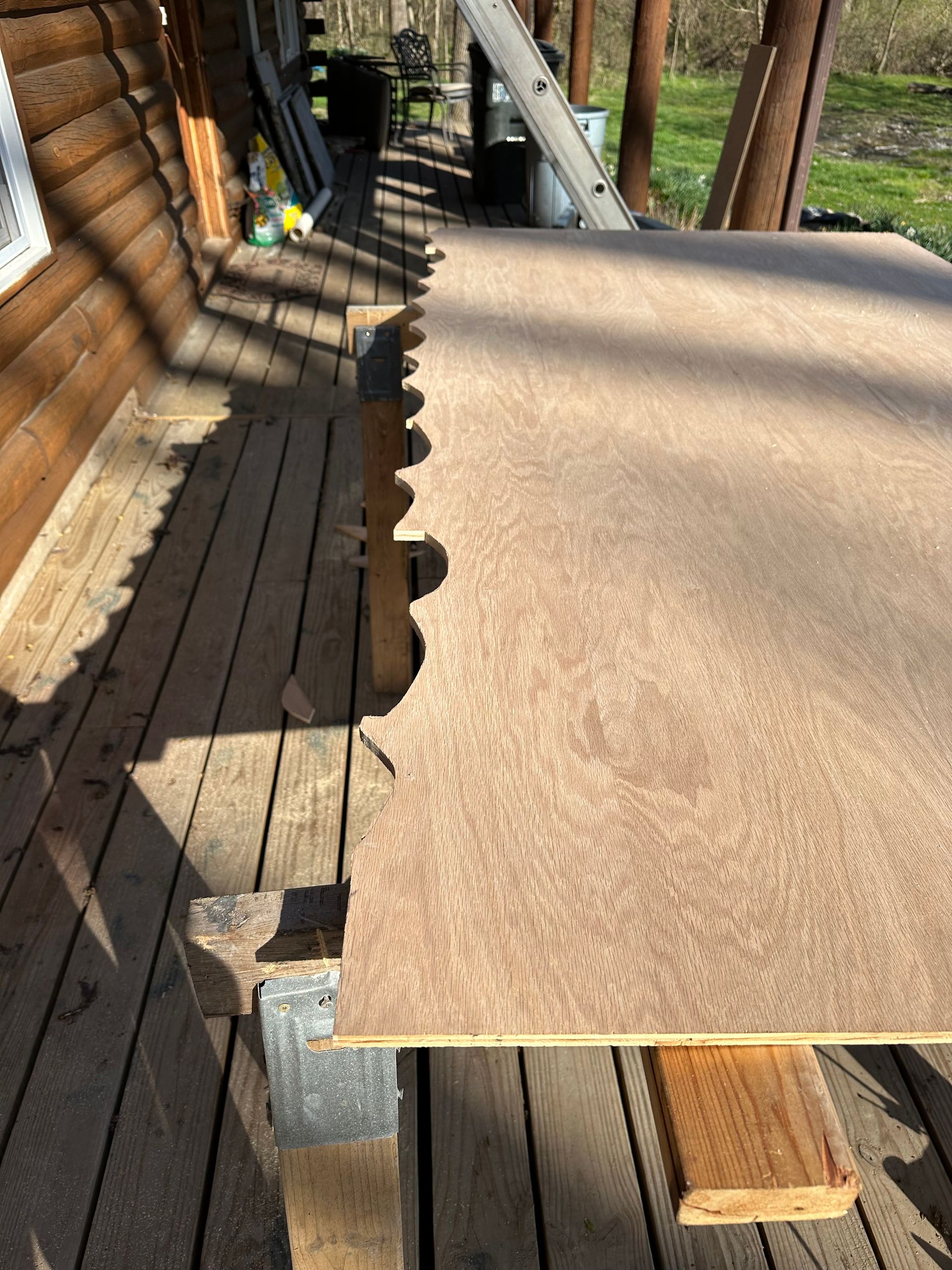 A large piece of plywood with a decorative edge sits on a sawhorse outside a log cabin, with sunlight casting shadows.