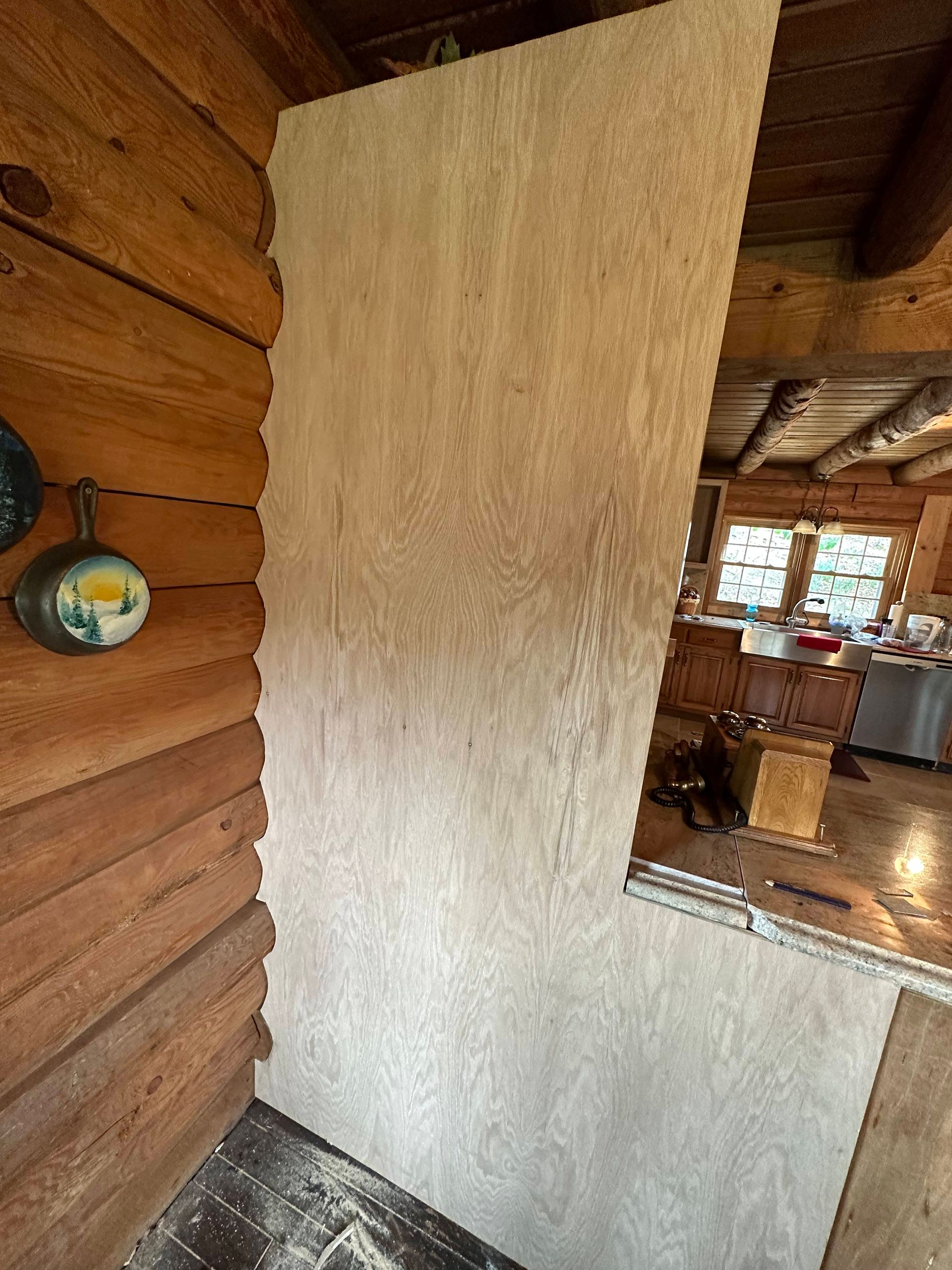 A plywood board is attached to a log cabin wall, likely for construction. Inside the cabin, a room and sunlight are visible.