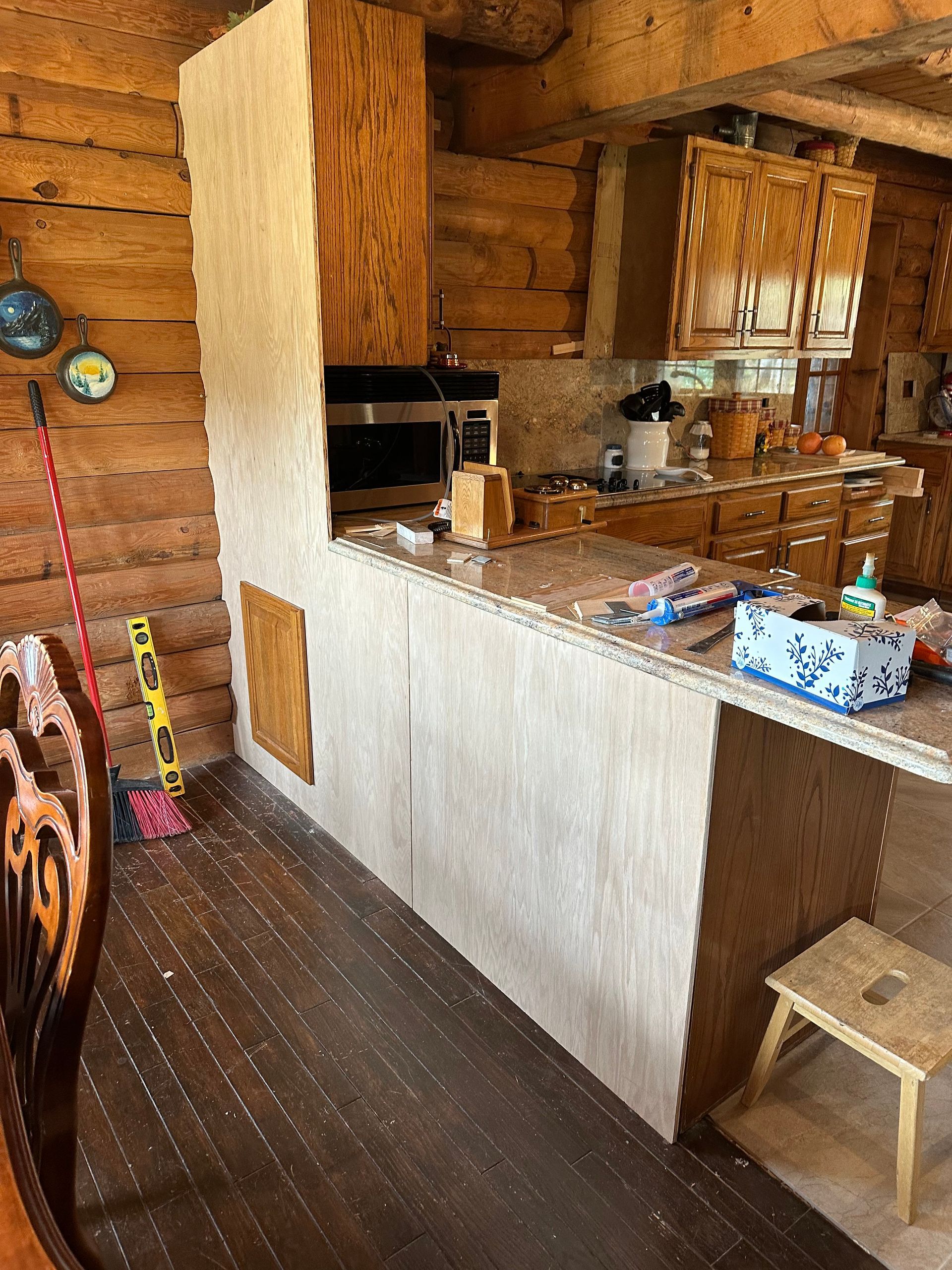 A rustic kitchen with wooden cabinets and a tall, light-colored countertop. Dark wood floors and a small stool are visible.