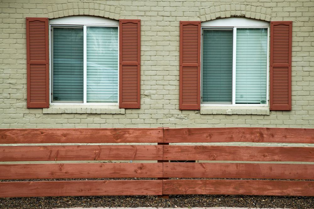 A Brick Building With Red Shutters And A Wooden Fence — Roo’s Door & Window Maintenance In Sippy Downs, QLD