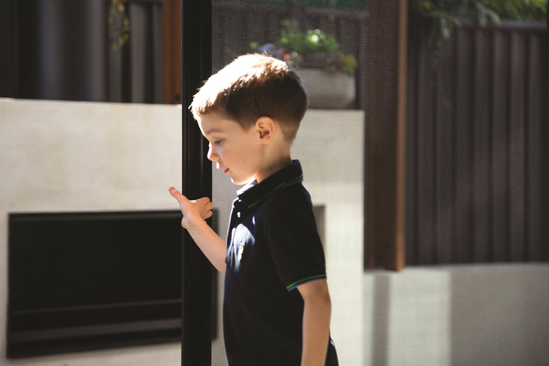 A Young Boy In A Black Shirt Is Standing In Front Of A Window — Roo’s Door & Window Maintenance In Buderim, QLD