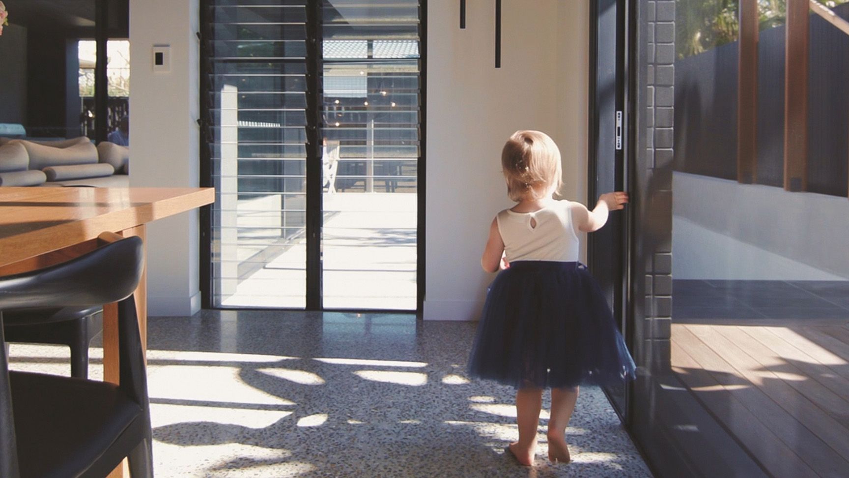 A Little Girl Is Standing In A Living Room Looking Out A Window — Roo’s Door & Window Maintenance In Sippy Downs, QLD