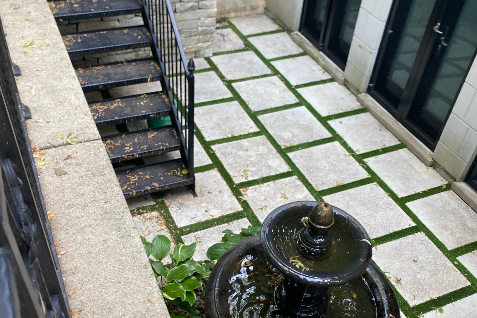 Exterior courtyard with stone pavers, grass in between, black stairs, and a fountain.