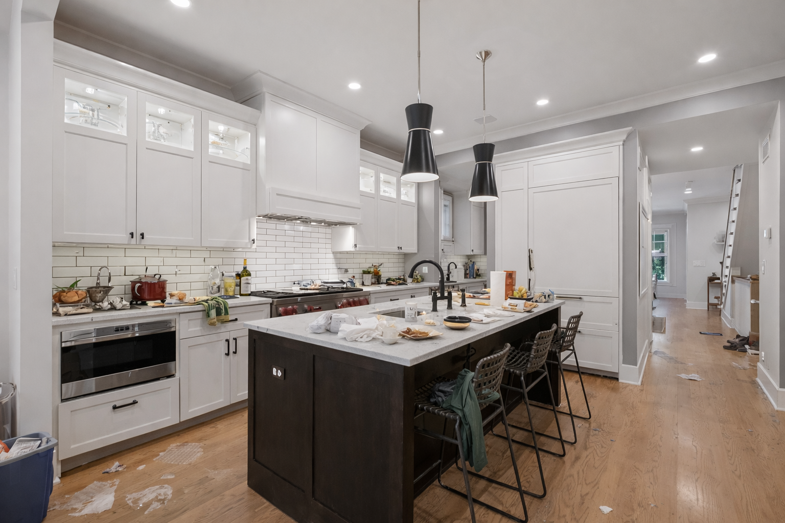 White kitchen with dark island and stools, messy countertops, two black pendant lights, and hardwood floors.
