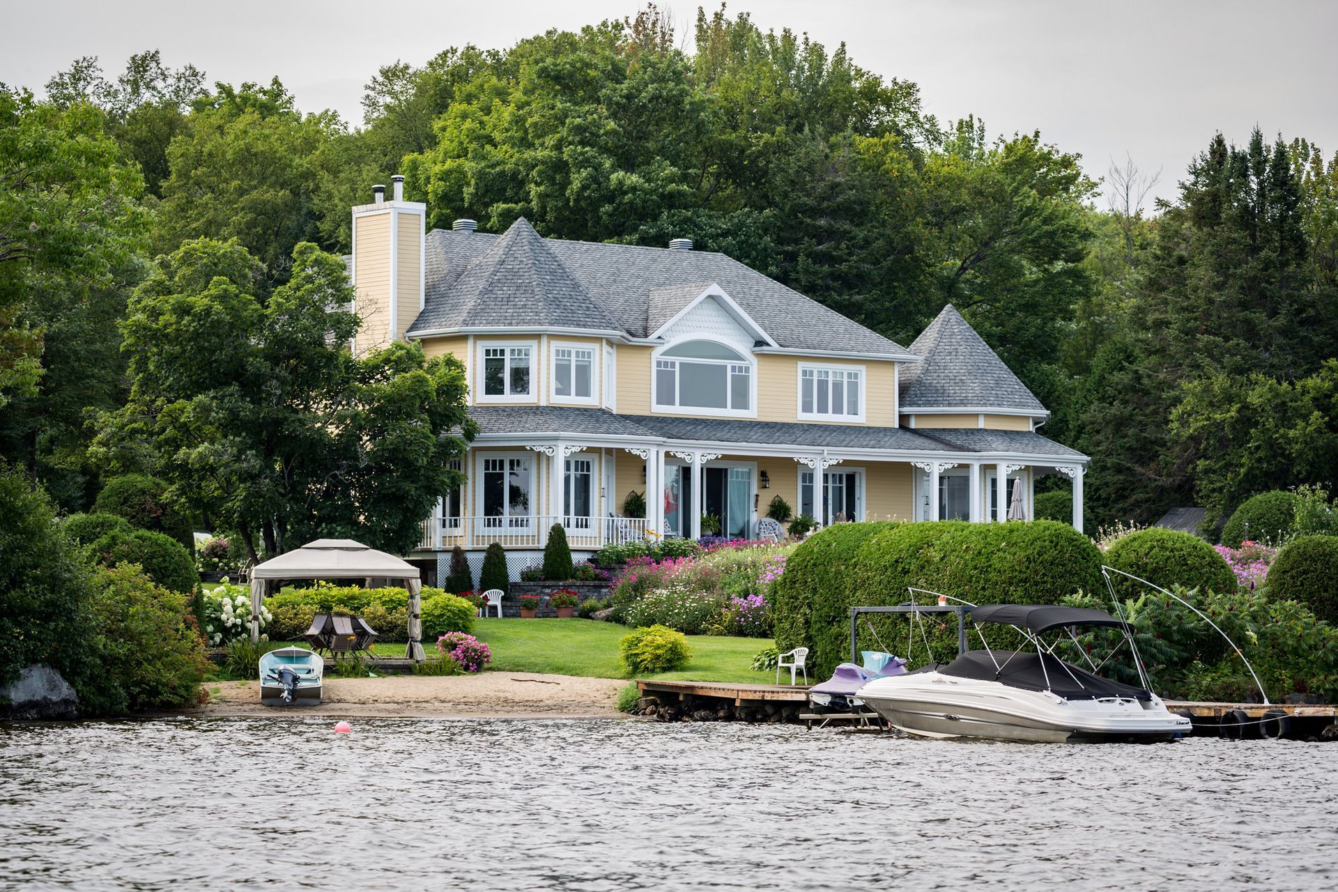 Yellow house with a lake view, boat, and gazebo. Lush green trees surround the house with a grey roof.