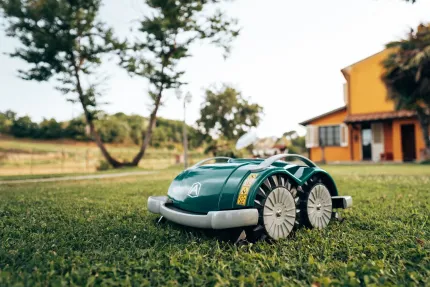 Green robotic lawnmower on grass in front of a yellow house, trees, and a field.