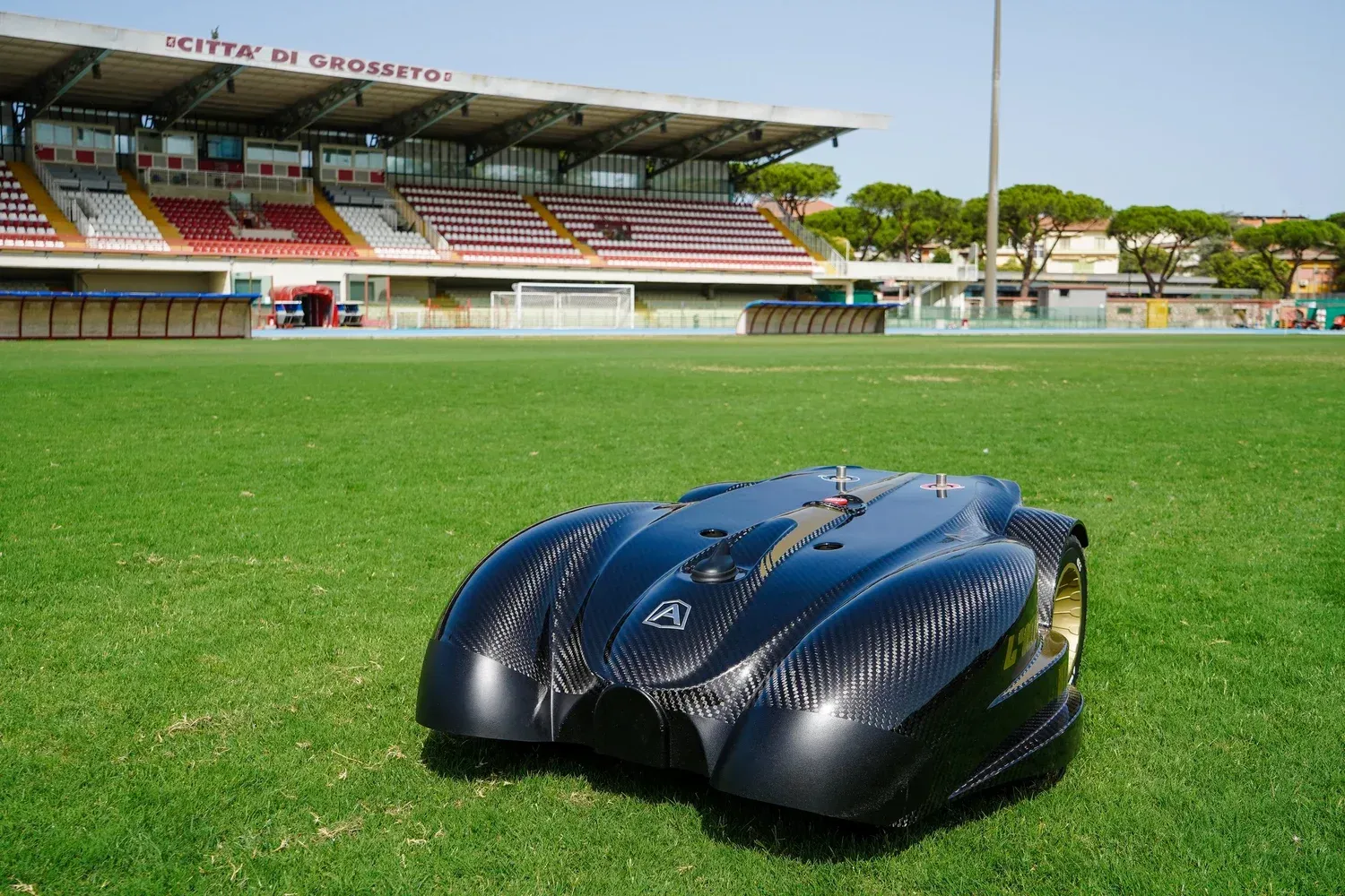 Robotic lawnmower on a green soccer field with stadium seating in the background.
