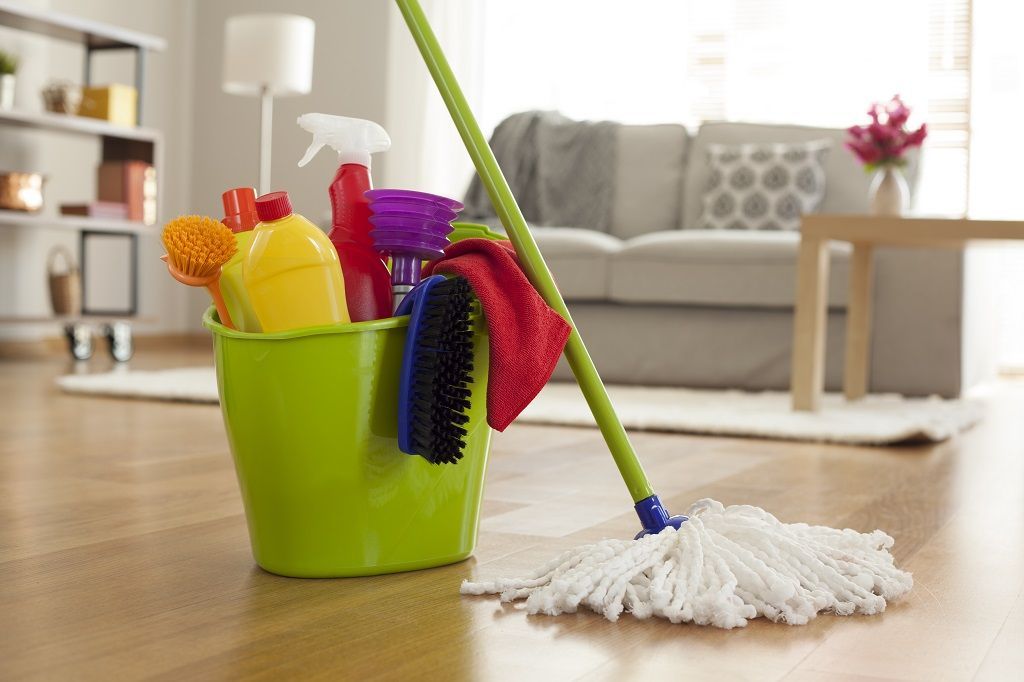 Cleaning supplies in a green bucket and a mop on a wooden floor in a living room.