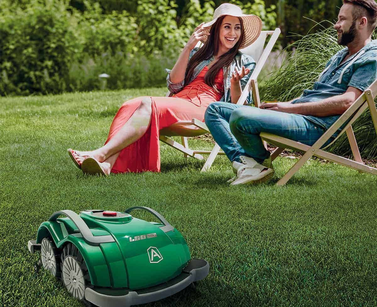 Robotic lawnmower cutting grass as couple relaxes in lawn chairs on sunny day.