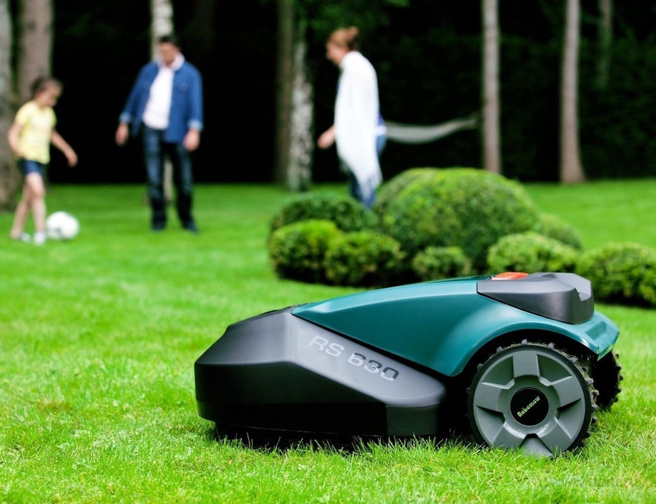 Robot lawnmower in a yard, family playing soccer in background.