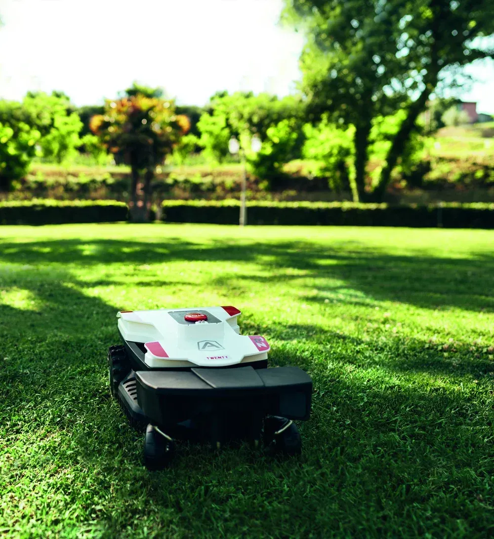 Robotic lawnmower on a green lawn, with trees and a hedge in the background.