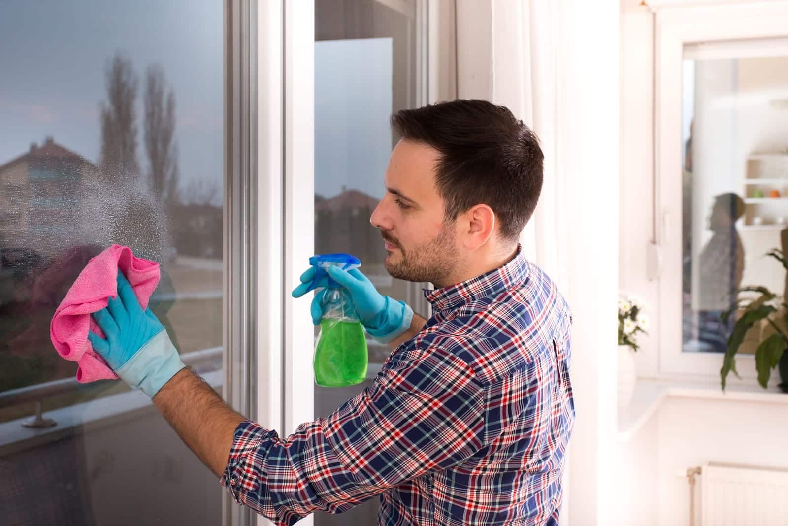 Man in plaid shirt and gloves sprays and wipes a window.