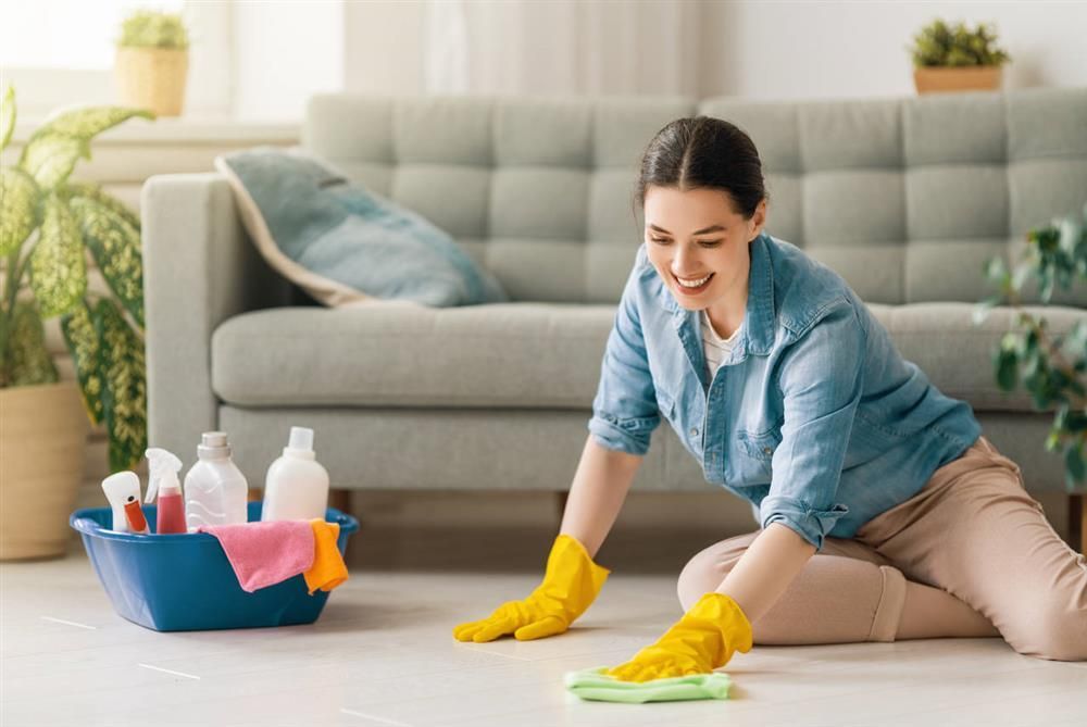 Woman wearing yellow gloves cleaning floor in a living room, smiling. Cleaning supplies in a blue bin.