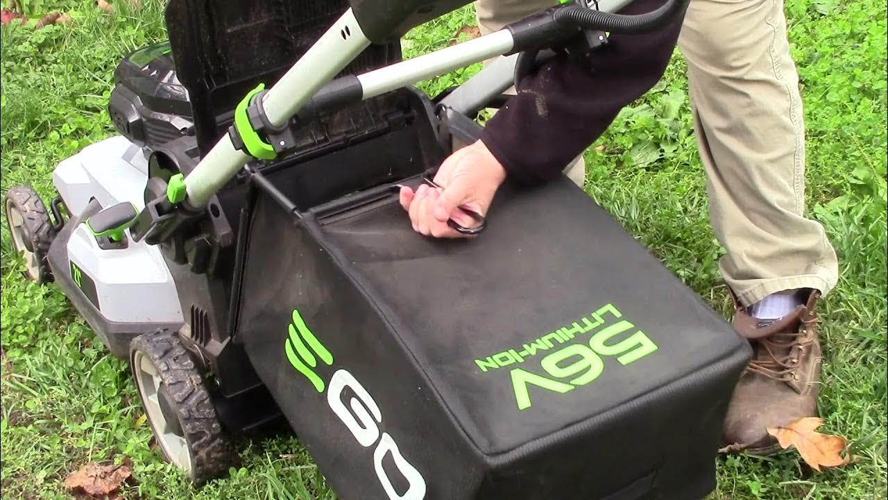 Person attaching a black grass collection bag to an electric lawnmower on green grass.