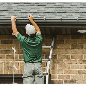 Person on a ladder hanging lights from a roof's gutter. A brick wall and gray roof are in the background.