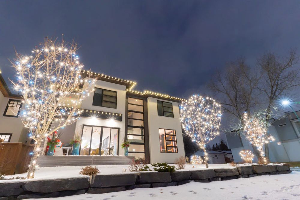 Two-story house with string lights on trees and roof at night. Snowy yard, blue and white lights.