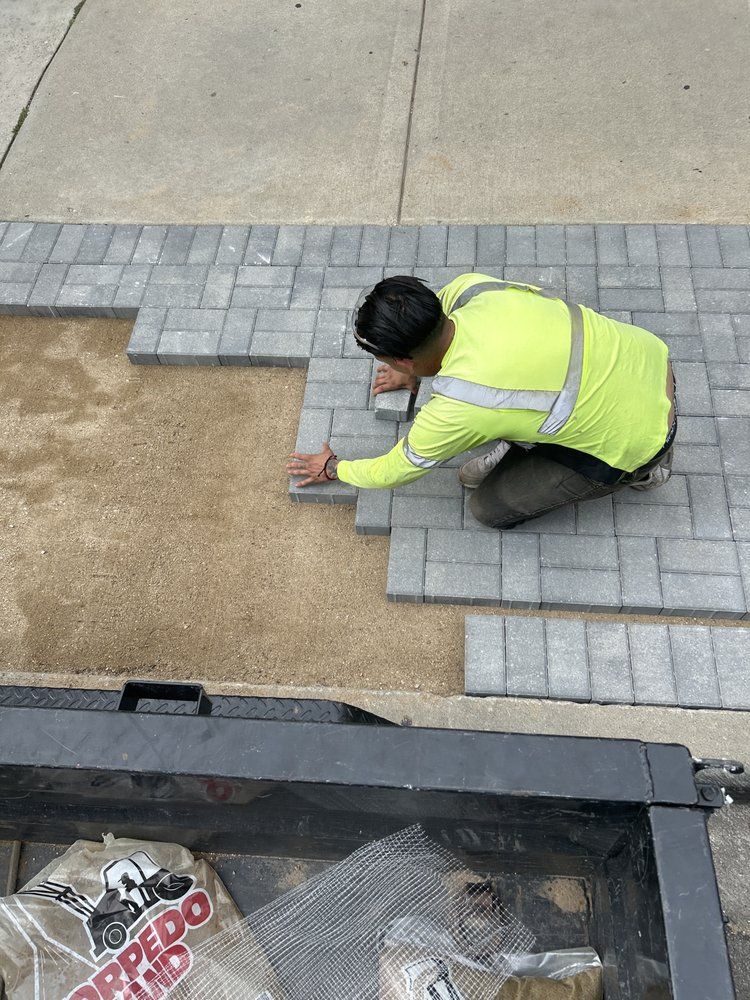 Person laying gray paving stones on a sandy base, wearing a yellow safety vest.