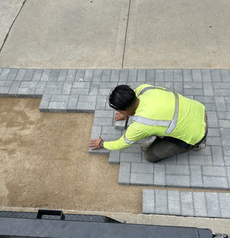 Person laying gray brick pavers on a sand base next to a concrete surface, wearing a neon yellow long-sleeve shirt.